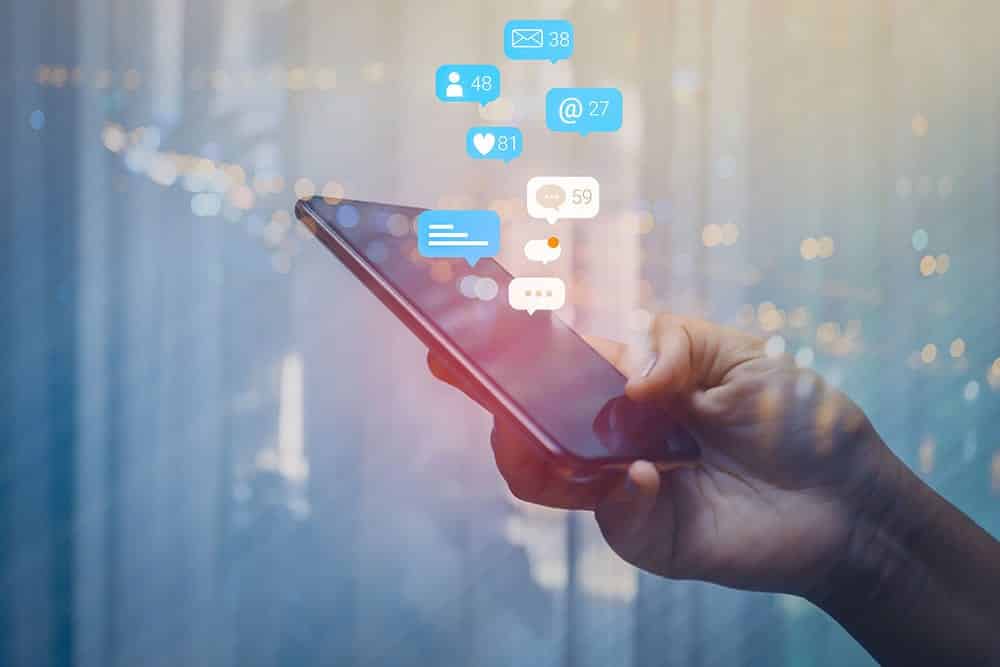 A hand holding a smartphone with social media icons coming out of it, representing the services of a social media manager.